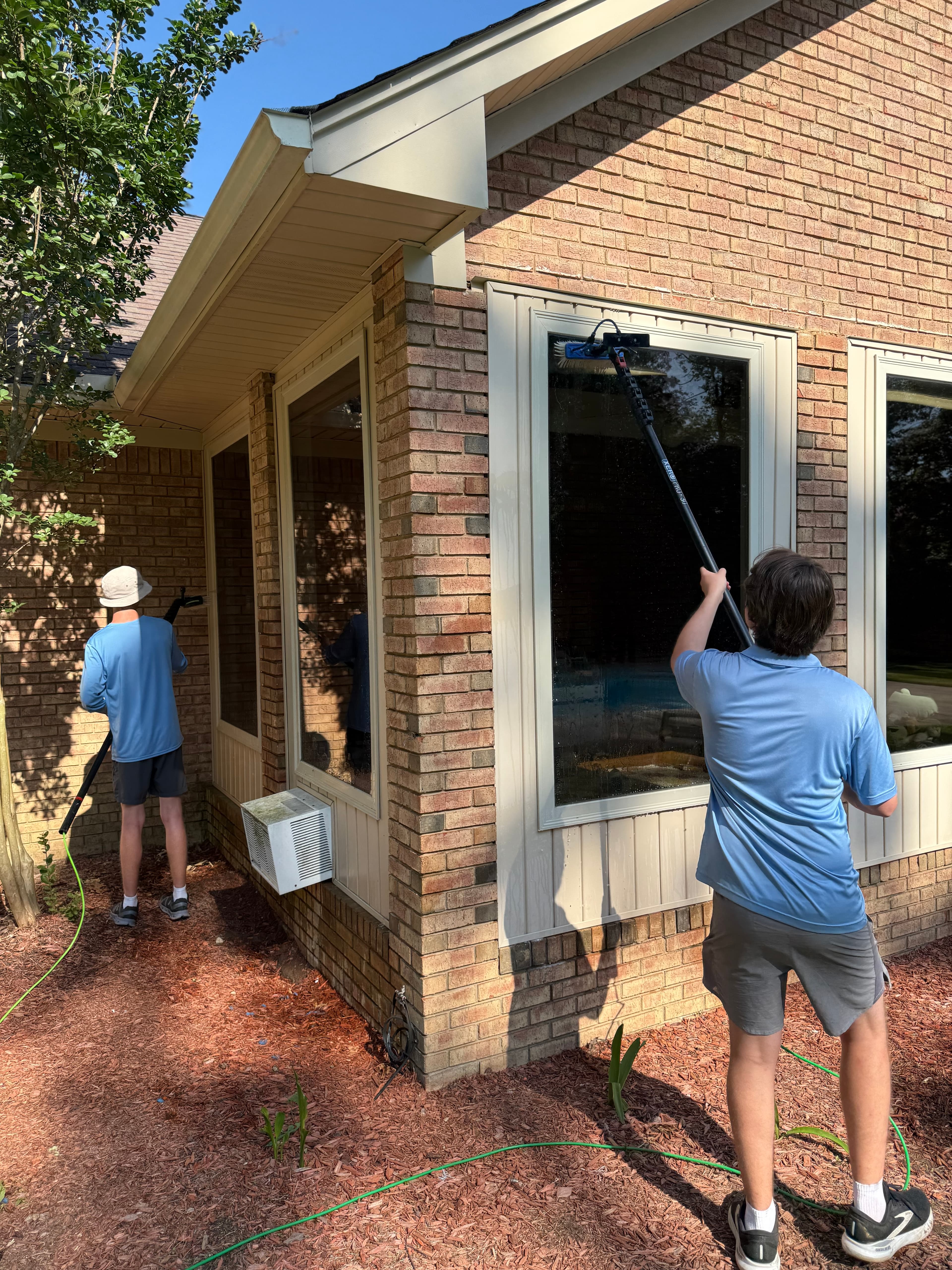 Two men cleaning large windows on a brick house using long-handled water-fed brushes.