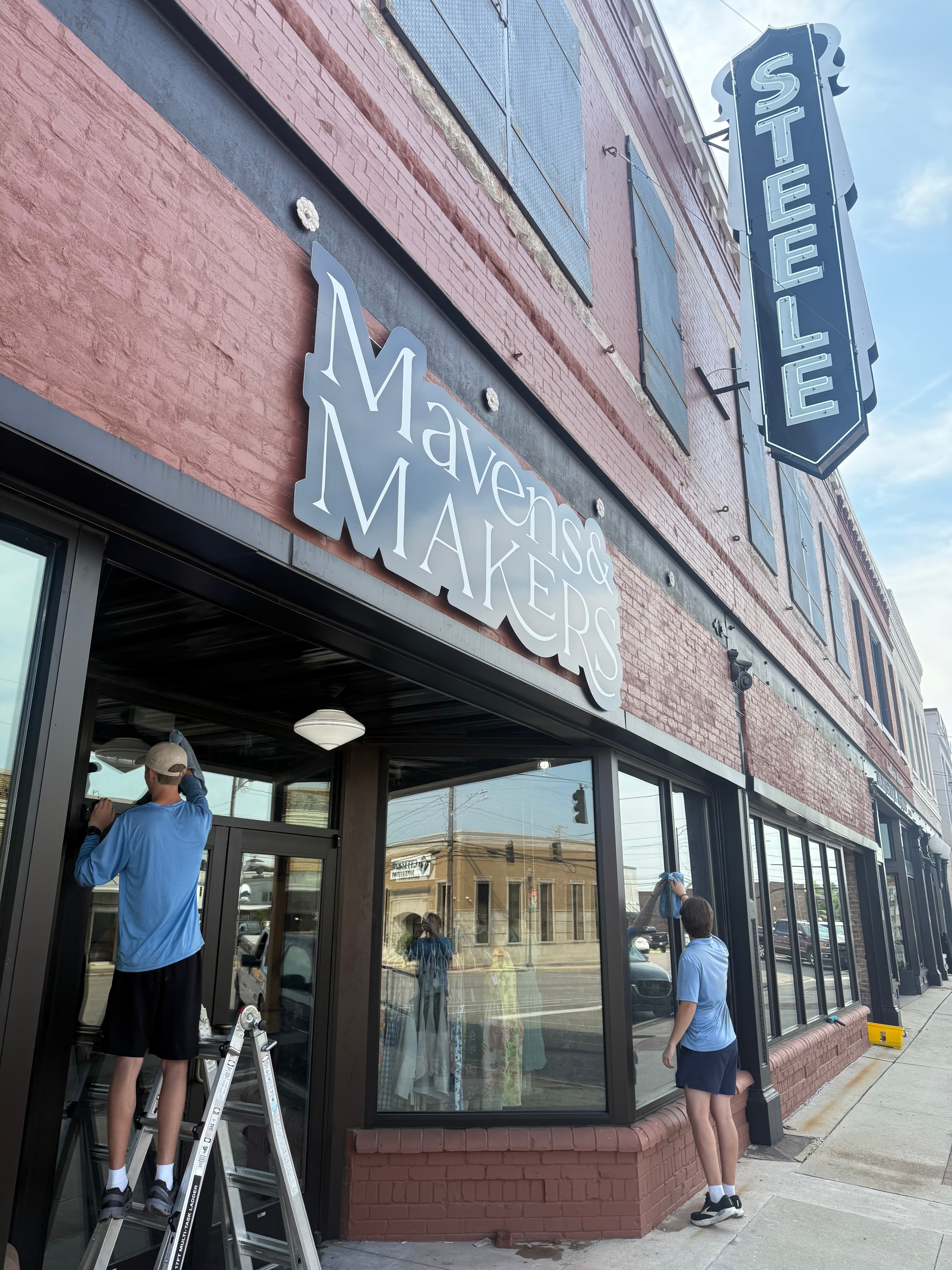 Workers cleaning windows of the Mavens & Makers storefront beneath a vintage Steele sign.