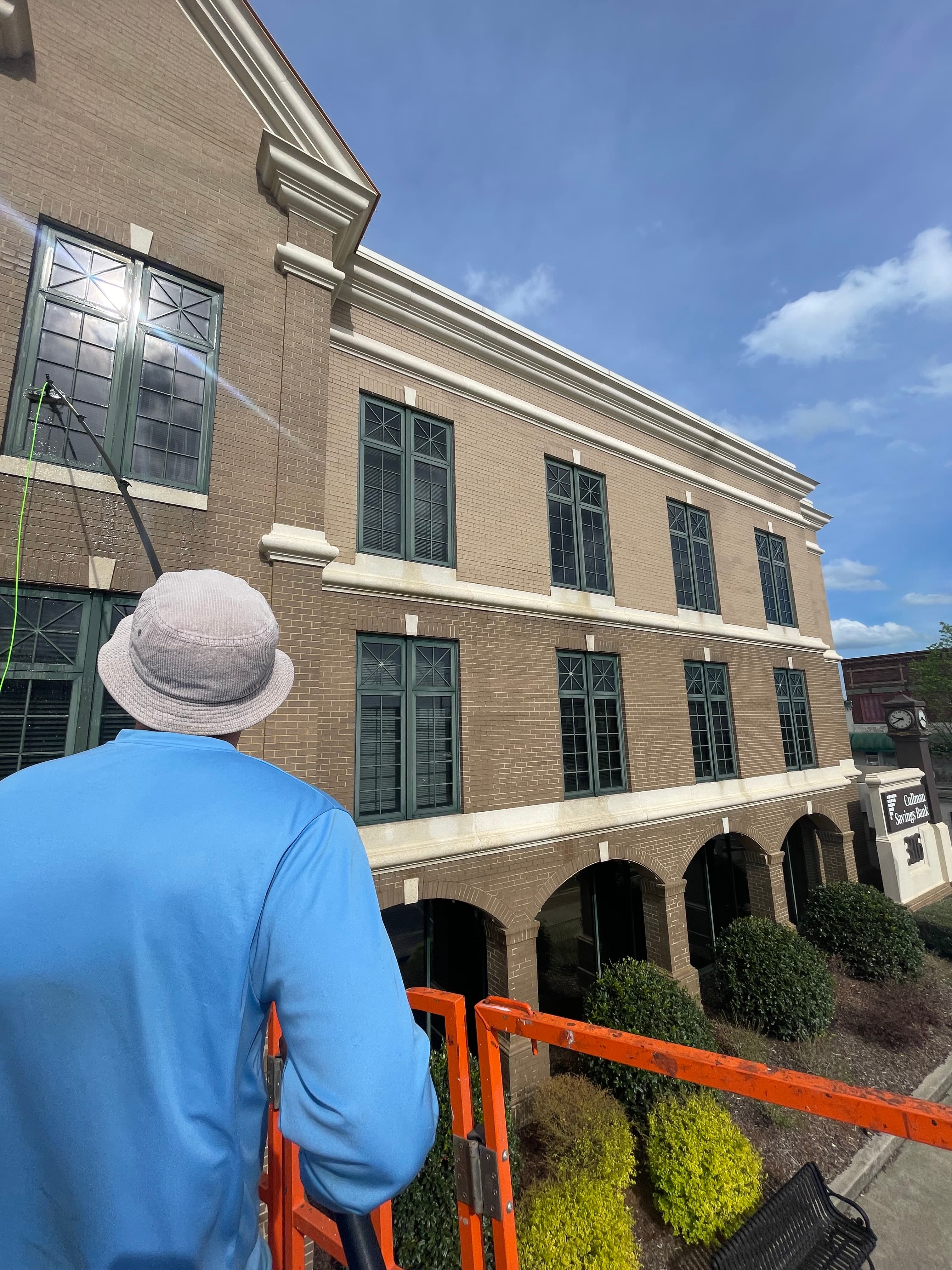 Worker on an orange lift cleaning windows of a brick building with a long pole.