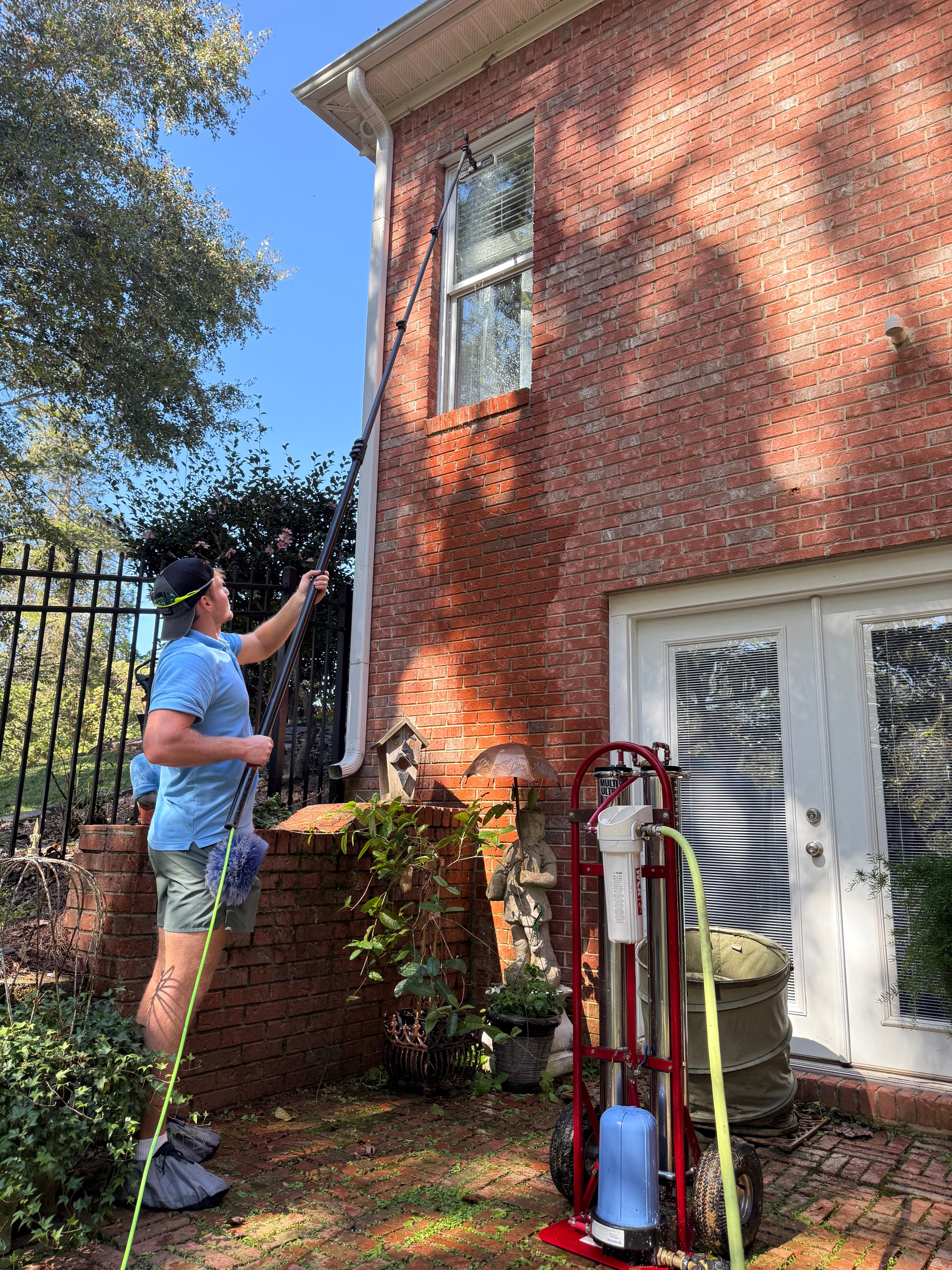 Man cleaning a window on a brick house with a long pole and filtration cart.