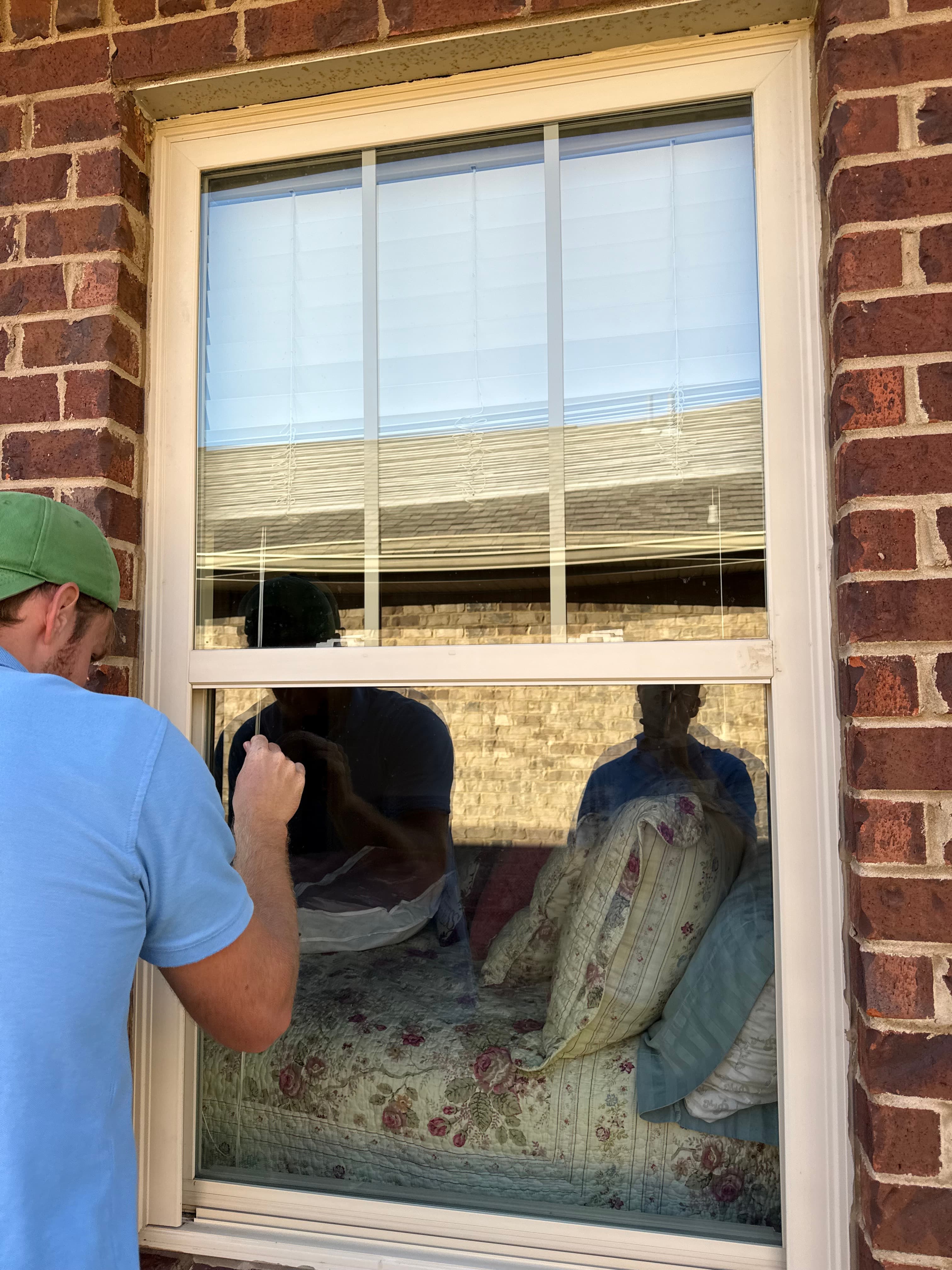 Man in a green cap looks through a window into a bedroom with floral bedding.