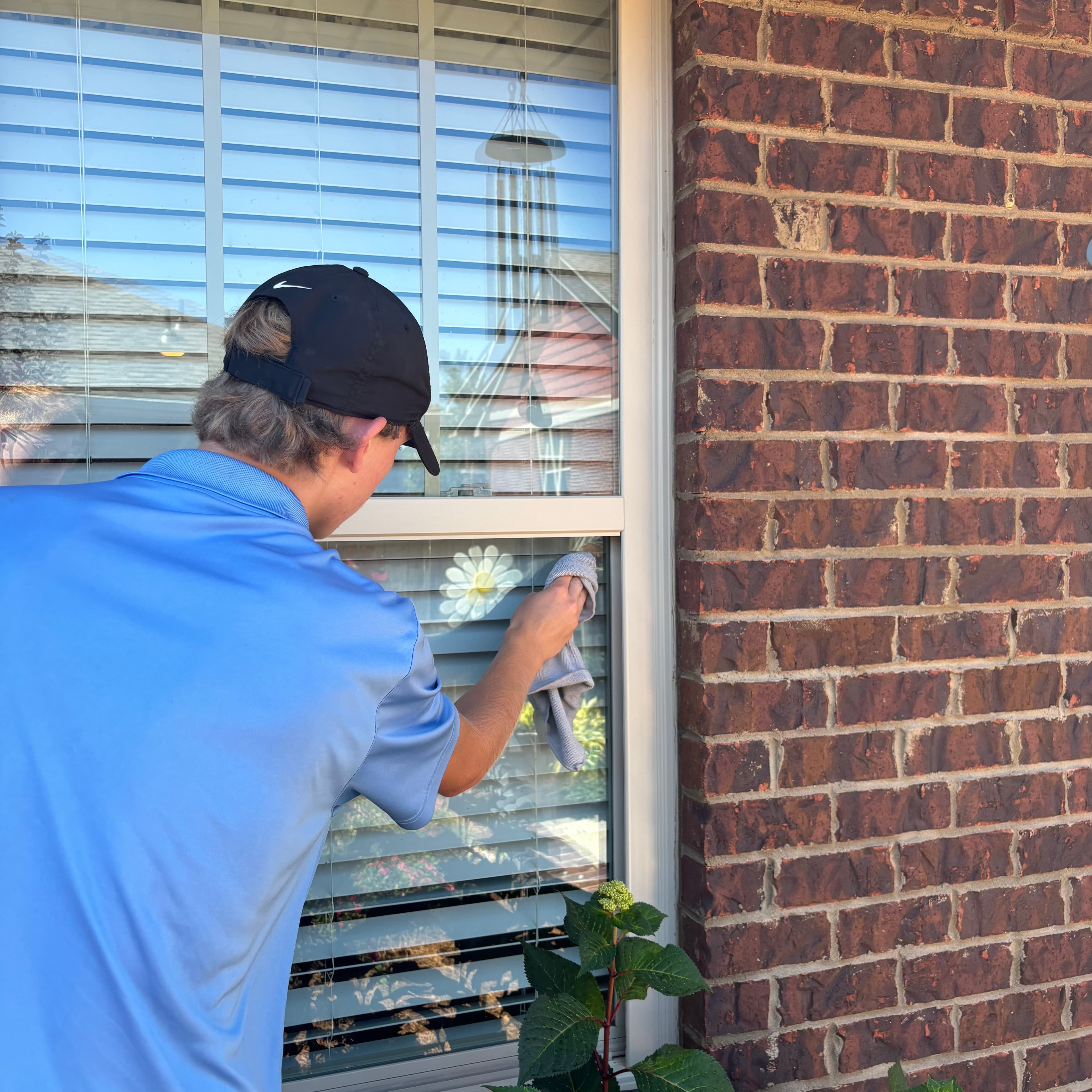 Man in blue shirt and black cap wipes a window on a brick house.