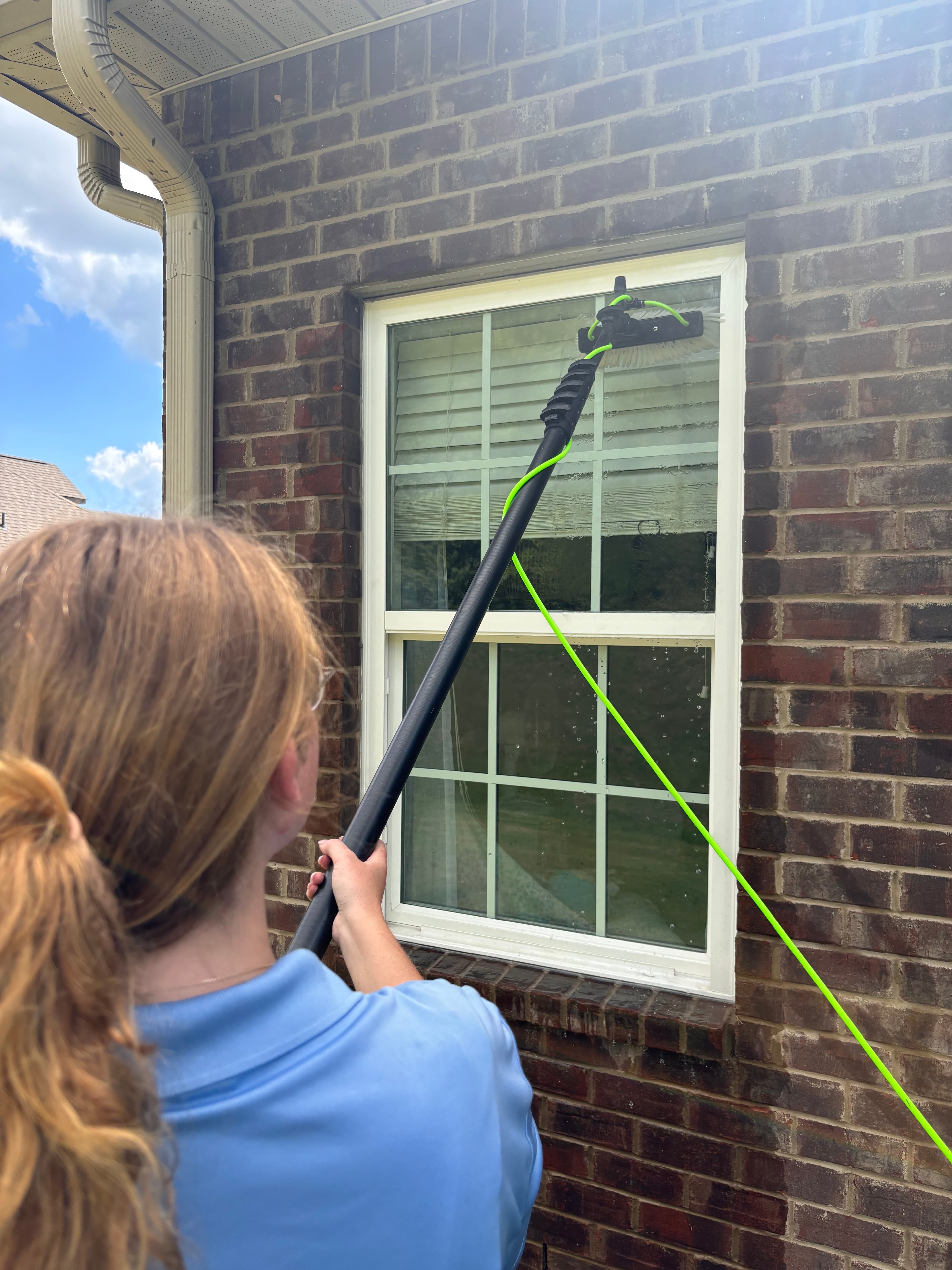 Person cleaning an exterior window with a long telescopic brush against a brick wall.