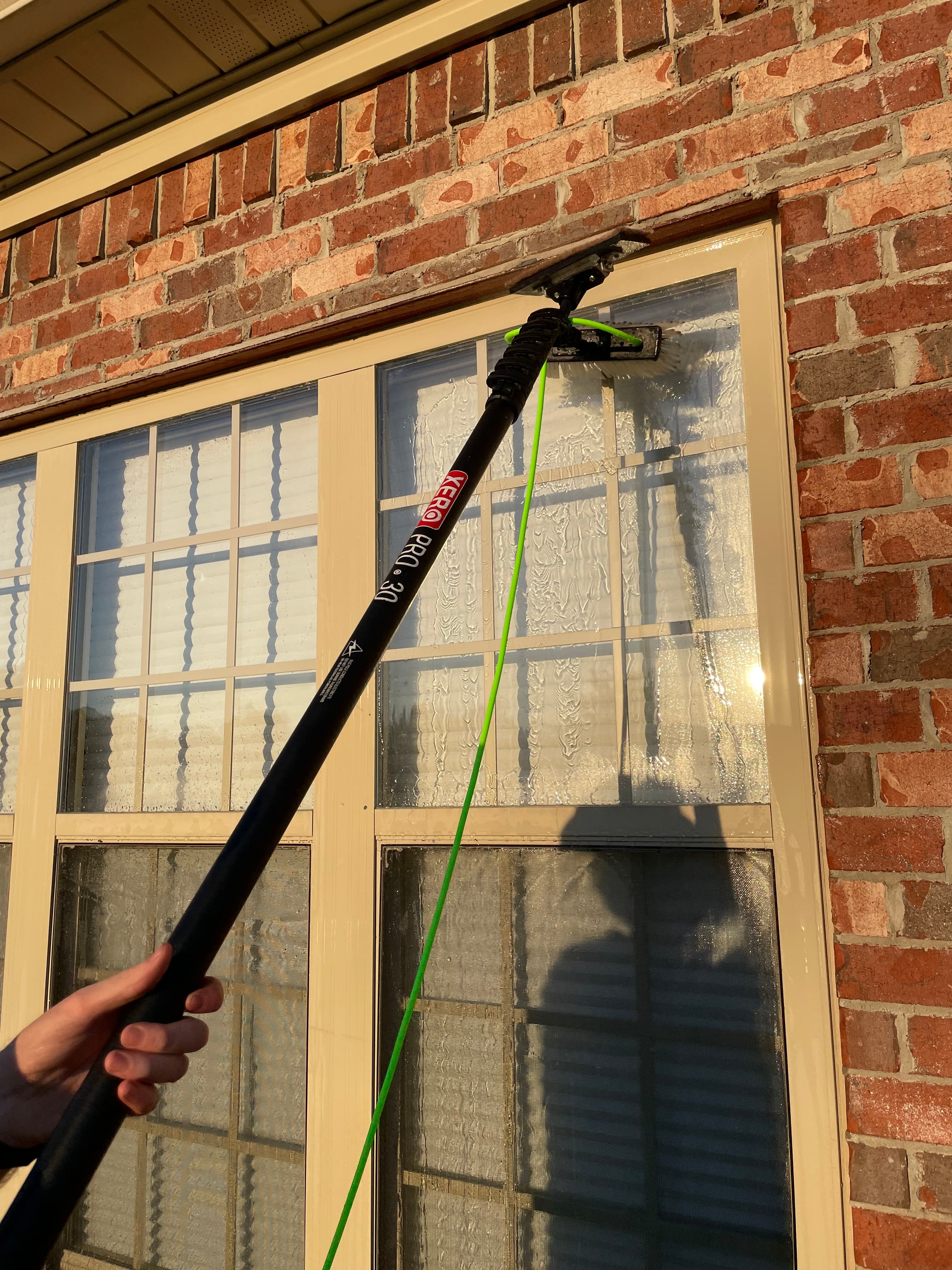 Hand using a water-fed pole to scrub a window on a red brick wall.