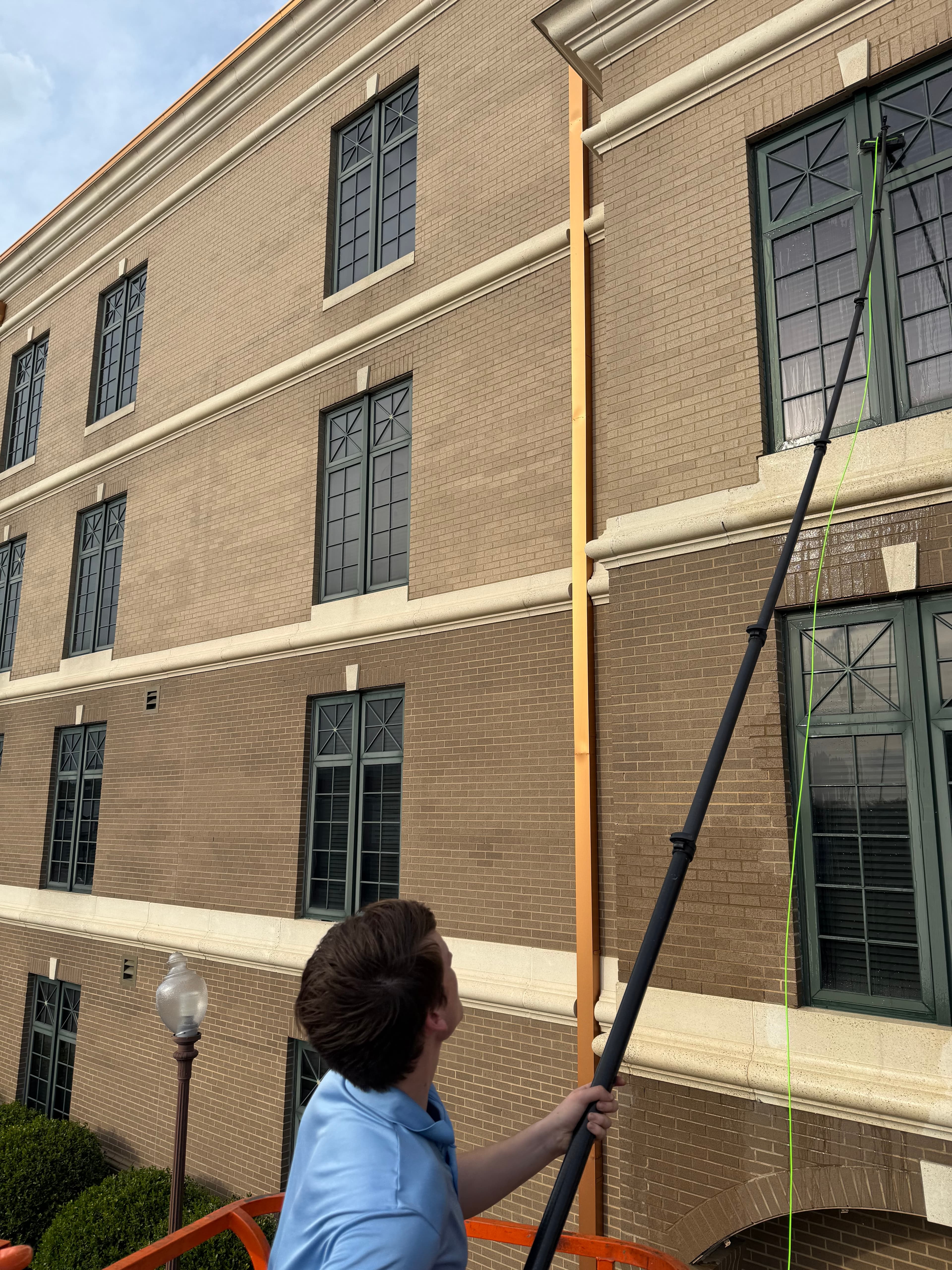 Man using a long telescopic pole to clean windows on a multi-story brick building.