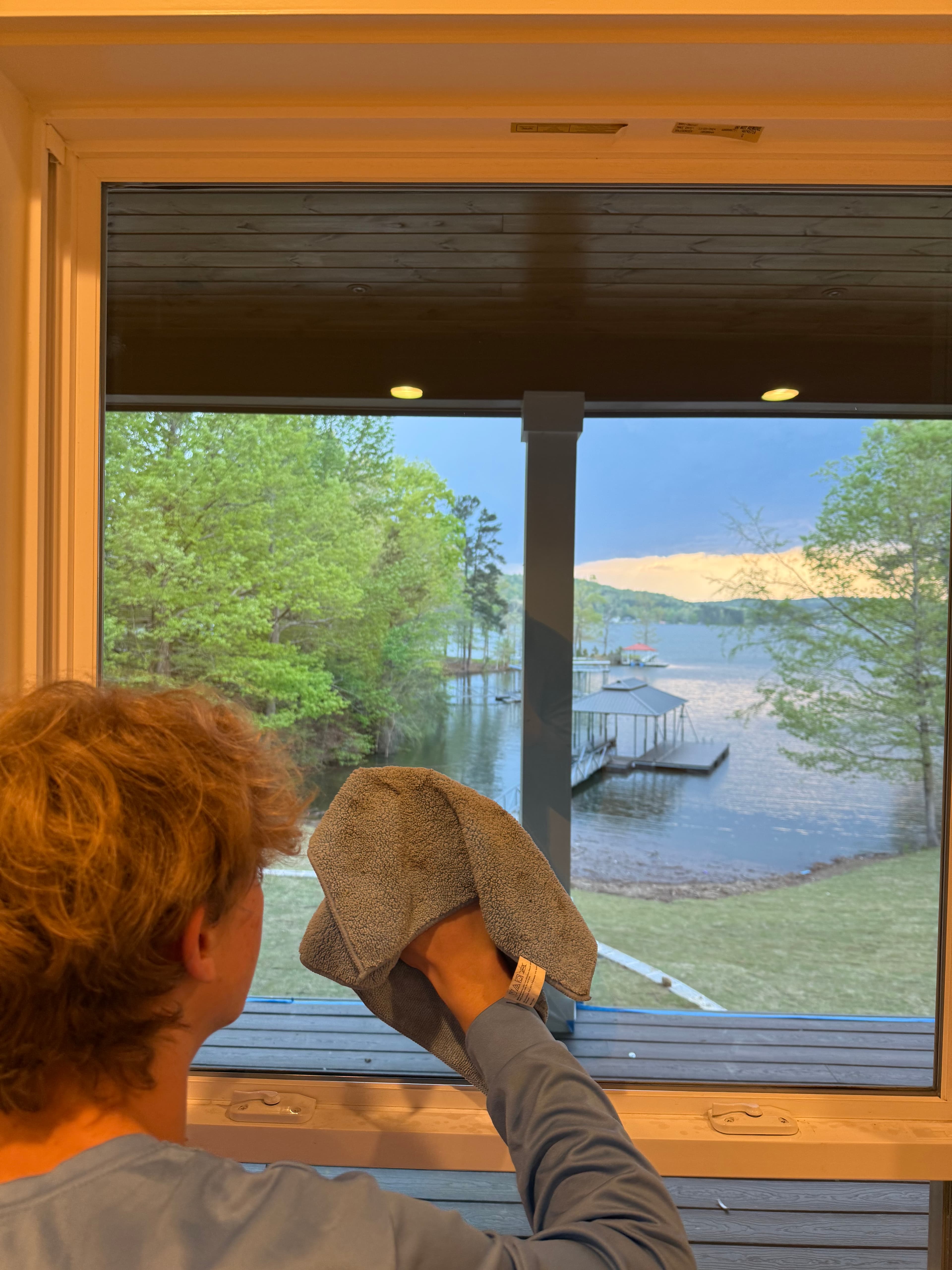 Person cleaning a window with a blue cloth, overlooking a scenic lake and green trees.